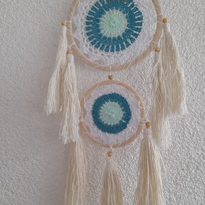 May include: Two white crocheted dreamcatchers with blue and green accents hanging from a white wall. The dreamcatchers have wooden hoops and white tassels.