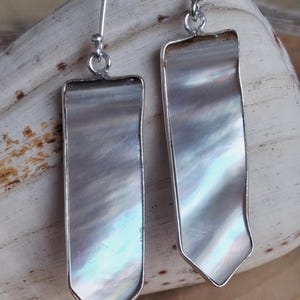 May include: A pair of rectangular drop earrings with a mother-of-pearl inlay and silver-colored metal trim. The earrings have a hook closure and a pointed bottom edge. The iridescent surface reflects light, creating a shimmering effect.