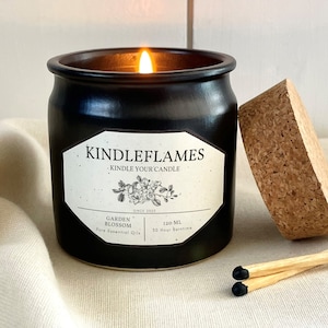 Op de afbeelding: Een zwarte keramische kaars pot met een kurken deksel en twee lucifers. De kaars is aangestoken en het etiket luidt "KINDLEFLAMES KINDLE YOUR CANDLE SINCE 2020 GARDEN BLOSSOM Pure Essential Oils 120 ml 50 Hour Burn Time".