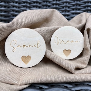 Könnte beinhalten: Zwei runde Holzscheiben mit den Namen "Samuel" und "Mama" in kursiv geschriebener Schrift. Jede Scheibe hat eine kleine Herzform, die in der Mitte ausgeschnitten ist.