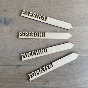 Könnte beinhalten: Vier hölzerne Pflanzenmarkierer mit den Worten "Paprika", "Pepperoni", "Zucchini" und "Tomaten" darauf.