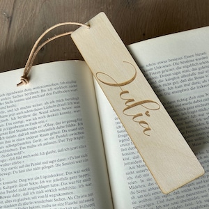 Könnte beinhalten: Ein Holz-Lesezeichen mit dem Namen "Julia" in kursiv geschriebener Schrift. Das Lesezeichen ist hellbraun und hat eine natürliche Holzmaserung.