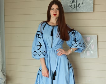 Könnte beinhalten: Ein hellblaues knielanges Kleid mit schwarzer Stickerei von Schwalben und geometrischen Rautenmustern an den bauschigen Ärmeln und am Rock. Das Kleid verfügt über einen passenden Bindegürtel in der Taille und schwarze Quasten am Vorderteil.