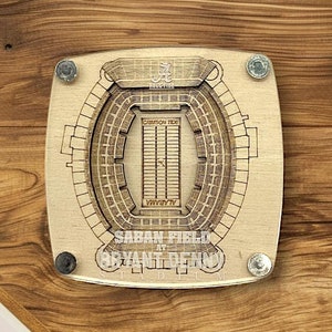 May include: Wooden coaster featuring a detailed outline of Bryant-Denny Stadium, home of the Alabama Crimson Tide. The coaster is made of wood and has a brown finish. The stadium is labeled "Saban Field at Bryant-Denny Stadium".
