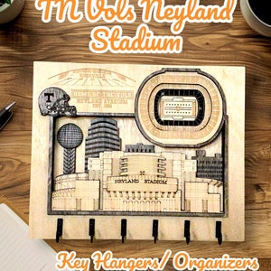 May include: Wooden key hanger featuring a detailed laser-cut depiction of Neyland Stadium, home of the Tennessee Volunteers. Includes a football helmet, stadium outline, and the text "TN Vols Neyland Stadium." Features five black key hooks.