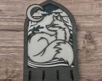 May include: A wooden key holder with a cutout design of a fox curled up under a crescent moon. The fox is white and the background is dark blue. There are four hooks on the bottom for hanging keys.
