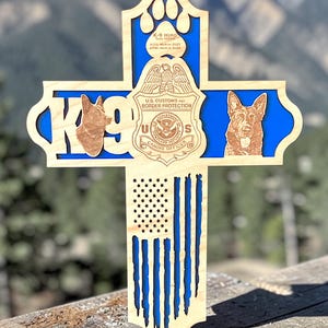May include: Wooden cross-shaped memorial with a blue background and an American flag design. Features the text "K-9" and images of dogs, along with the text "U.S. Customs and Border Protection Canine Officer". Includes the name "K-9 Hiro".