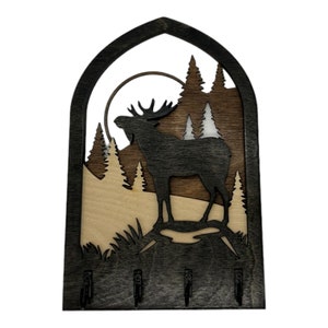 May include: Wooden key holder with a silhouette of a moose standing in a forest scene. The key holder has four hooks and is painted in shades of brown and black.