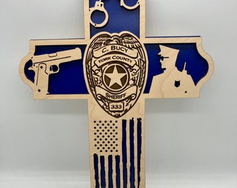 Op de afbeelding: Houten kruis met een blauwe achtergrond en een sheriff-badge met de tekst "C. BUCY YORK COUNTY SHERIFF 333". Het kruis heeft ook een pistool, handboeien, een silhouet van een politieagent en een Amerikaanse vlag.
