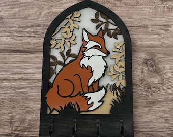May include: A wooden key holder with a fox silhouette in a forest scene. The fox is orange and white with a black nose and eyes. The forest scene is made up of brown and beige leaves and branches. The key holder has three hooks.
