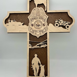 May include: A wooden cross with a brown stain. The cross features a Border Patrol badge, a horse and rider, a helicopter, a person with a dog, a boat, and an ATV. The badge reads "Border Patrol" and "U.S. Patrol Agent".