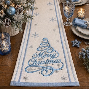 May include: Cream-colored Christmas table runner with a blue border and embroidered Christmas tree design. The words "Merry Christmas" are embroidered in blue. The runner is decorated with snowflakes.