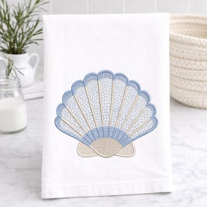 Może przedstawiać: Biały ręcznik z haftowanym wzorem muszli. Muszla jest szczegółowo wykonana z jasnoniebieskiej i beżowej nici, tworząc teksturowaną, nadmorską estetykę. Ręcznik jest złożony i prezentowany na białej powierzchni.