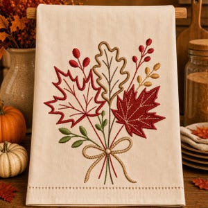 May include: A cream-colored kitchen towel featuring an embroidered bouquet of autumn leaves, including red maple and tan oak leaves, accented with red berries and green sprigs. The design is tied with a tan embroidered bow at the base.