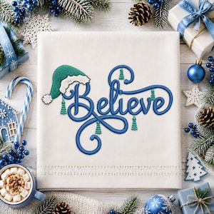 Può includere: Un quadrato di tessuto bianco con un ricamo in blu e verde. La parola Believe è scritta in corsivo blu, con la lettera B sormontata da un cappello da Babbo Natale verde. Piccole nappe verdi pendono dalle lettere blu.