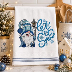 May include: A white kitchen towel featuring an embroidered gnome wearing a blue patterned hat and holding a lantern. The text "let it snow" is embroidered in blue script next to the gnome, accompanied by several blue snowflake designs.