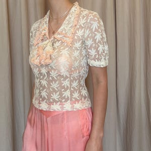 Pu&ograve; includere: Top in pizzo color crema vintage con maniche corte e delicato motivo floreale. Il top presenta un bordo arricciato color pesca intorno allo scollo e un accento color pesca abbinato sul petto. Abbinato a pantaloni color pesca a vita alta e gamba larga.