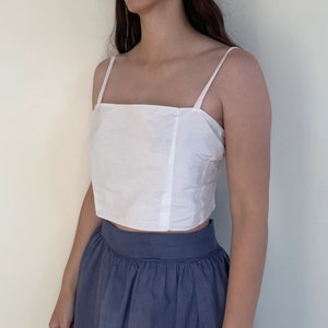 Può includere: Un top bianco corto con spalline sottili e scollo quadrato. Il top è abbinato a una gonna blu scuro.