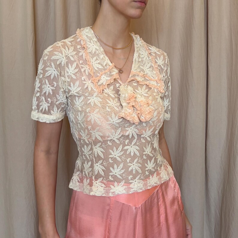 Pu&ograve; includere: Un top in pizzo color crema con maniche corte e motivo floreale. Il top ha un colletto a balze color pesca e un accento abbinato allo scollo. Abbinato a una gonna rosa chiaro.