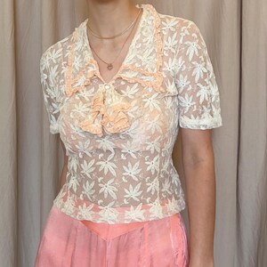 Pu&ograve; includere: Un top in pizzo color crema con un motivo floreale. Il top presenta maniche corte, un colletto a balze e dettagli color pesca sul colletto e sul petto. &Egrave; abbinato a pantaloni rosa.