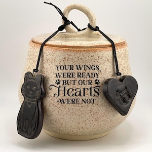 Puede incluir: Una urna para mascotas de cerámica moteada con tapa y asa. La urna tiene un gato negro y un dije en forma de corazón colgando. El texto en la urna dice "YOUR WINGS WERE READY BUT OUR HEARTS WERE NOT."