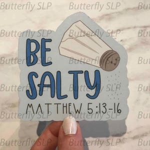 May include: A light blue sticker with the words "BE SALTY" in blue, with a salt shaker illustration. The text "MATTHEW 5:13-16" is below. The sticker is held by a person's hand.