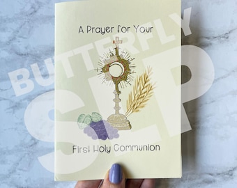 May include: A small, white booklet with a yellow background. The front cover features a drawing of a golden monstrance with a white host, wheat stalks, and grapes. The text on the cover reads "A Prayer for Your First Holy Communion".