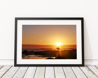 May include: A framed photograph of a silhouette of a surfer walking on a beach at sunset. The sun is setting over the ocean, casting a warm glow on the scene.