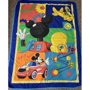 Puede incluir: Una manta rectangular con un estampado colorido de Disney que muestra a Mickey Mouse conduciendo un coche rojo, a Pluto tumbado y una gran silueta de Mickey Mouse. El diseño incluye formas geométricas, engranajes y el texto BEEP! BEEP! sobre un borde azul.