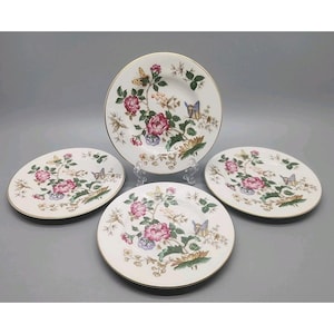 Puede incluir: Un juego de cuatro platos redondos de cerámica blanca con un patrón floral de rosas rosas, hojas verdes y mariposas coloridas. Cada plato tiene un borde dorado fino y un diseño botánico que incluye pequeñas flores blancas y tallos marrones.