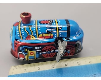 Pode incluir: Uma locomotiva de brinquedo de folha-de-flandres azul com mecanismo de corda, rodas vermelhas, uma pequena chaminé vermelha e detalhes em amarelo e preto. Uma chave de metal está fixada na lateral. O brinquedo está ao lado de uma régua de 9 cm.