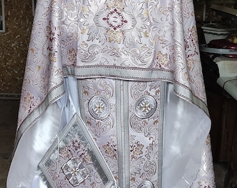 May include: A white and silver liturgical vestment with intricate floral embroidery and a silver trim. The vestment has a white sash and a small silver pouch with a tassel.
