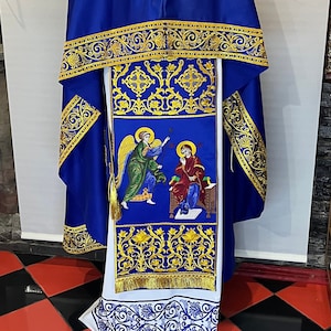 May include: A blue and white liturgical vestment with gold embroidery and a depiction of an angel and a figure in a red robe. The vestment is displayed on a mannequin against a checkered black and red floor.