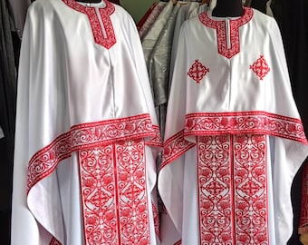 May include: Two white liturgical robes with red embroidered trim and floral patterns. The robes have a cross design on the chest and a wide band of red embroidery around the bottom.