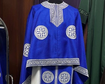 Könnte beinhalten: Ein blaues und weißes liturgisches Gewand mit silberner Stickerei. Das Gewand hat eine weiße Basis mit einem blauen Saum und eine blaue Kapuze mit silberner Stickerei. Die Stickerei zeigt Kreuze und andere geometrische Muster.