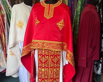 May include: A red liturgical vestment with gold embroidery. The vestment has a white undergarment and a long, flowing red stole with gold embroidery. The vestment is hanging on a rack in a store.