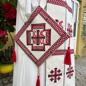 May include: White liturgical vestment with red and black embroidery. The vestment features a cross with the inscription "IC XC NIKA" and a red tassel.