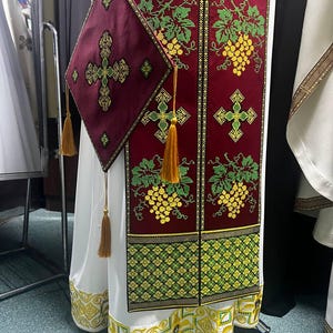 K&ouml;nnte beinhalten: Ein wei&szlig;es liturgisches Gewand mit einem roten und goldenen bestickten Feld mit Trauben und Kreuzen. Das Gewand hat eine passende rote und goldene bestickte Tasche mit einem Kreuz.