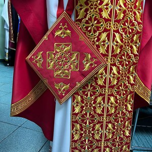 May include: Close-up of a red and gold embroidered religious garment. The garment features intricate gold floral and cross patterns on a red background. A matching square piece is also visible, along with a white undergarment.