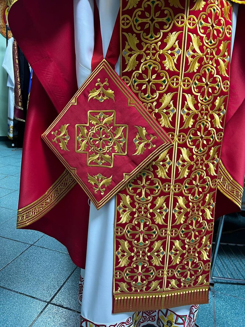 May include: Close-up of a red and gold embroidered religious garment. The garment features intricate gold floral and cross patterns on a red background. A matching square piece is also visible, along with a white undergarment.