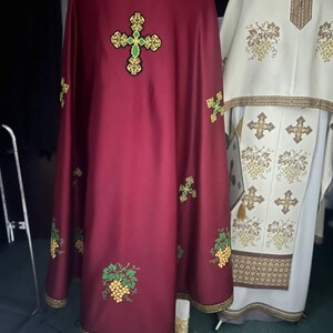 K&ouml;nnte beinhalten: Ein burgunderfarbenes liturgisches Gewand mit goldenen Verzierungen und gestickten Weinreben und Kreuzen. Das Gewand hat ein gro&szlig;es goldenes Kreuz mit gr&uuml;nem Zentrum auf der R&uuml;ckseite.