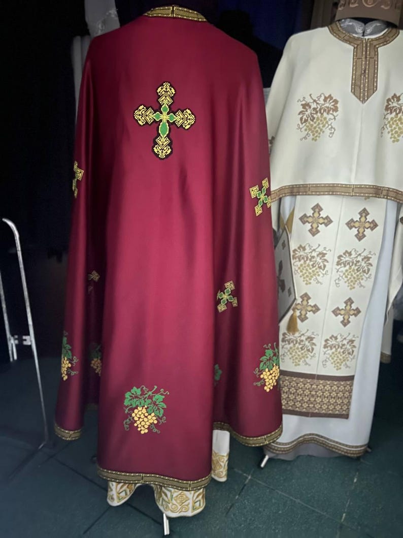K&ouml;nnte beinhalten: Ein burgunderfarbenes liturgisches Gewand mit goldenen Verzierungen und gestickten Weinreben und Kreuzen. Das Gewand hat ein gro&szlig;es goldenes Kreuz mit gr&uuml;nem Zentrum auf der R&uuml;ckseite.