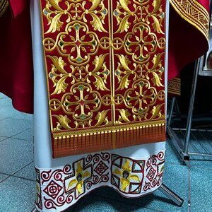 May include: A red and gold embroidered liturgical vestment. The vestment features intricate gold floral patterns on a red background. The bottom hem is white with a repeating pattern of red and gold designs, including winged figures. The vestment is displayed on a silver stand.
