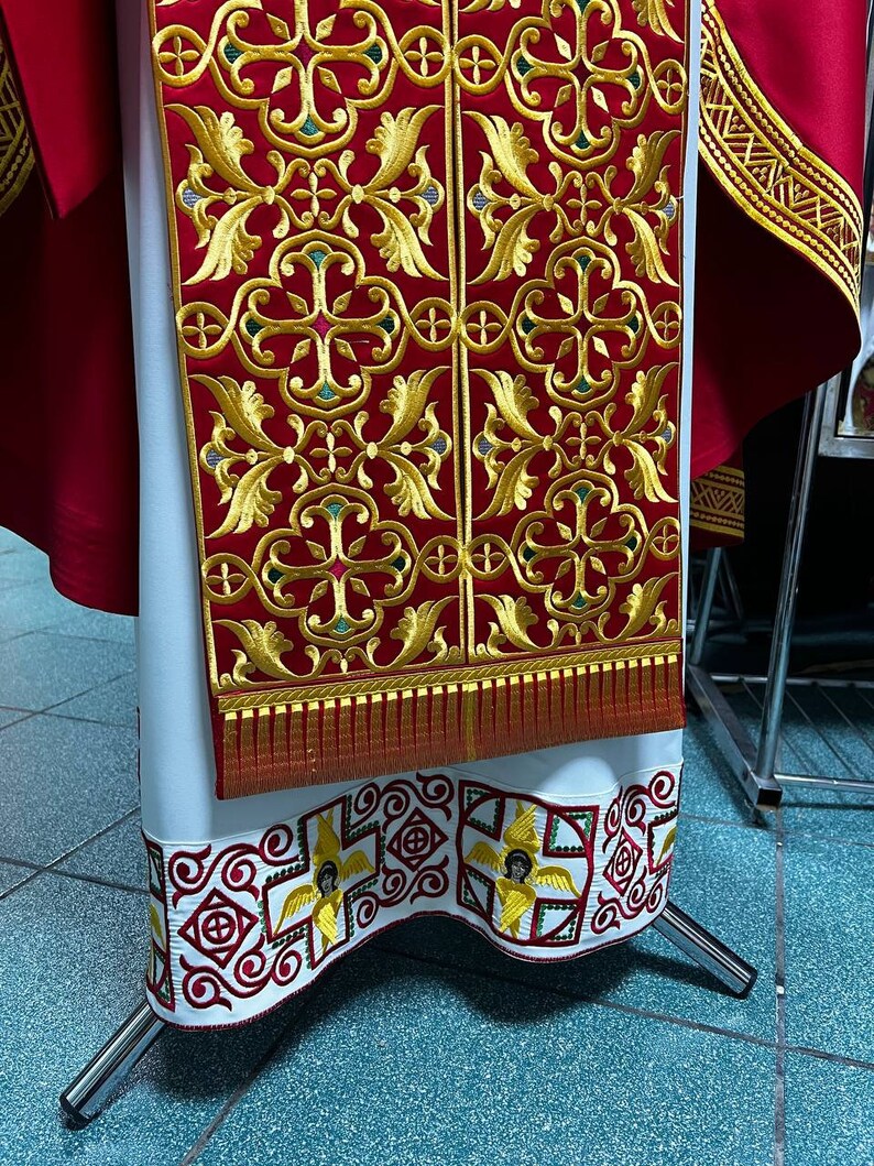 May include: A red and gold embroidered liturgical vestment. The vestment features intricate gold floral patterns on a red background. The bottom hem is white with a repeating pattern of red and gold designs, including winged figures. The vestment is displayed on a silver stand.