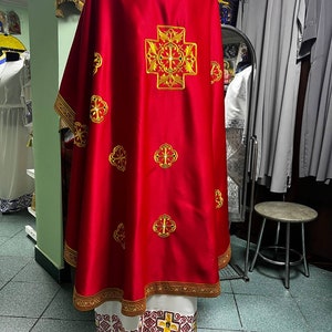 May include: A red liturgical vestment with gold embroidered crosses and trim. The back of the vestment features a large, ornate cross design. The bottom hem and edges are trimmed with gold and white patterns. The garment is displayed on a mannequin.