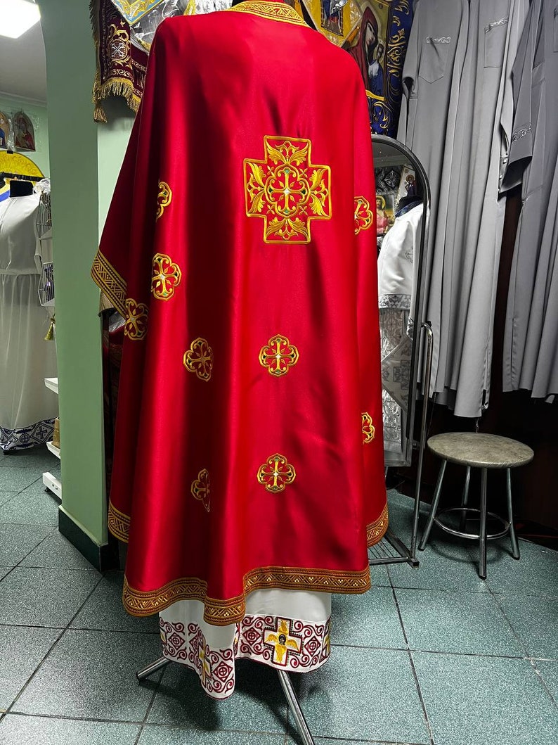 May include: A red liturgical vestment with gold embroidered crosses and trim. The back of the vestment features a large, ornate cross design. The bottom hem and edges are trimmed with gold and white patterns. The garment is displayed on a mannequin.