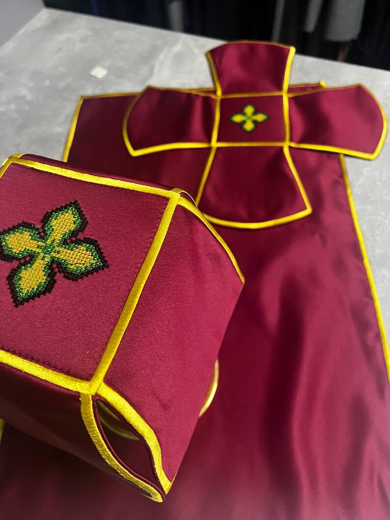 K&ouml;nnte beinhalten: Ein liturgisches Set in Burgunderrot mit goldenen Verzierungen und gestickten gr&uuml;nen und gelben Kreuzen. Das Set beinhaltet ein Kelchtuch und eine Burse.