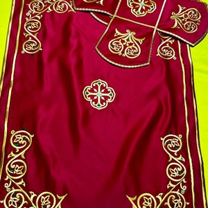 May include: A burgundy liturgical set featuring gold embroidered detailing. The set includes a rectangular cloth with corner designs and a central cross, along with a matching cross-shaped cover. The embroidery incorporates floral and geometric patterns.