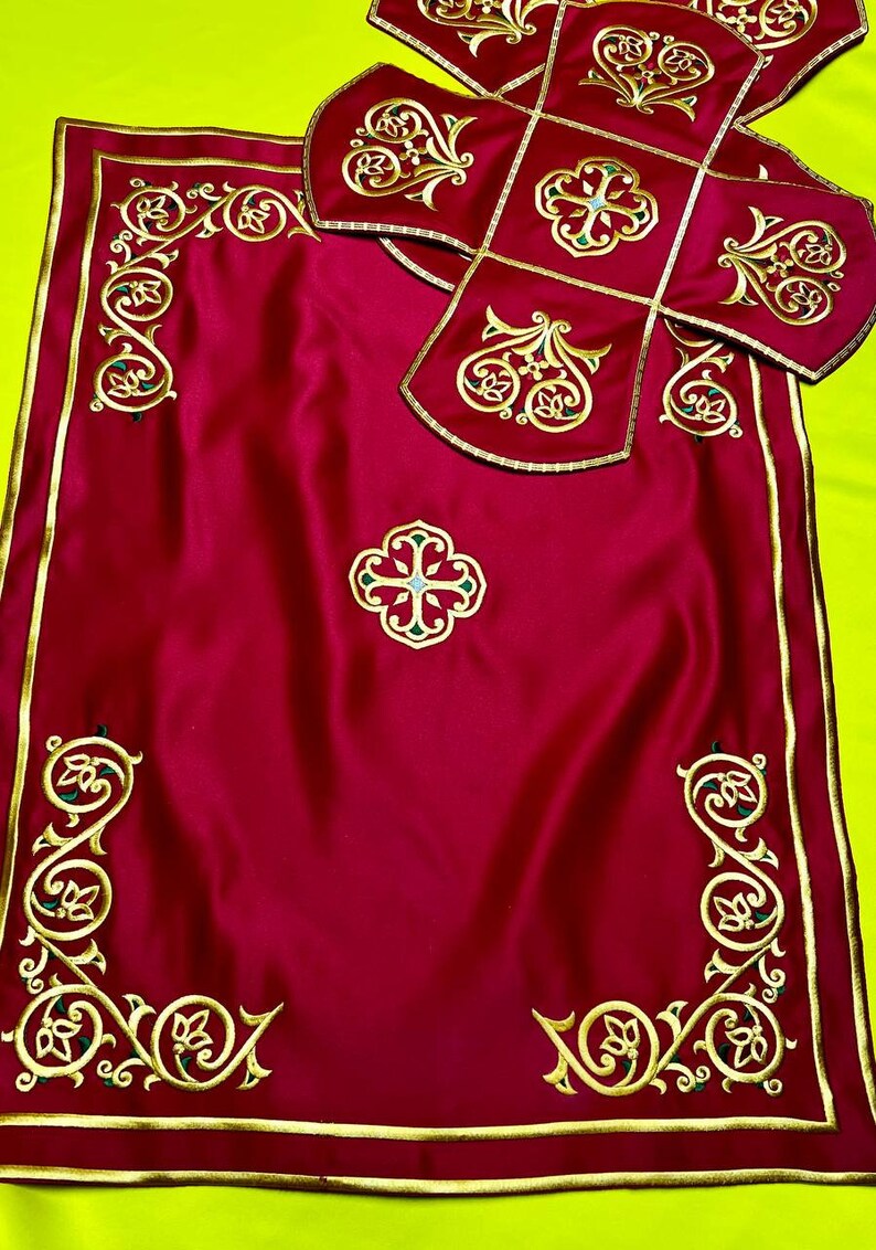 May include: A burgundy liturgical set featuring gold embroidered detailing. The set includes a rectangular cloth with corner designs and a central cross, along with a matching cross-shaped cover. The embroidery incorporates floral and geometric patterns.