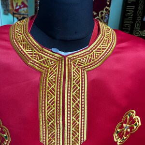 May include: A red liturgical vestment with gold embroidery. The vestment features a detailed gold embroidered collar and a central panel with vertical gold patterns. Decorative gold and green embroidered floral designs are on the lower part of the vestment.